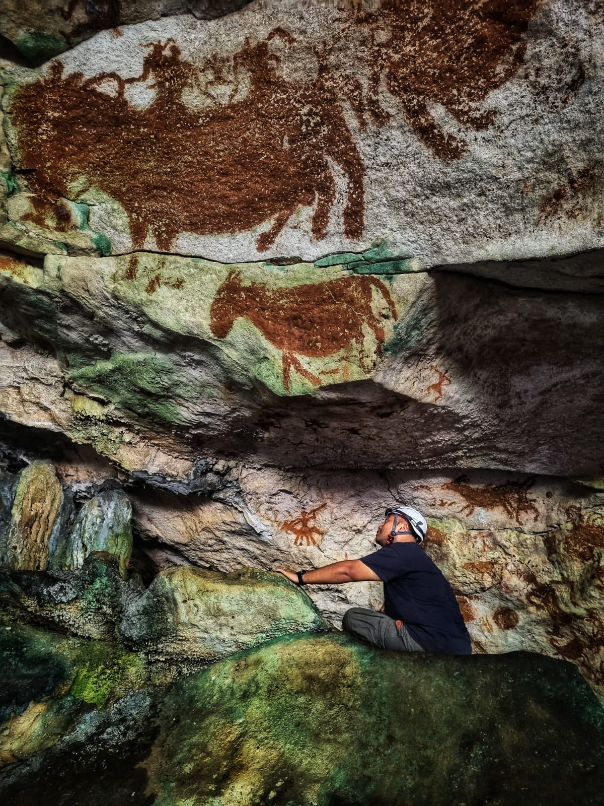 A researcher in cave with a head torch on crouches down, looking up at some red ochre rock art images of frankly very cute fat horses that get bigger, the higher up the wall they are painted. One horse has two human riders and tiny figure iseither pointing or aiming a weapon at another horse A researcher in cave with a head torch on crouches down, looking up at some red ochre rock art images of frankly very cute fat horses that get bigger, the higher up the wall they are painted. One horse has two human riders and tiny figure iseither pointing or aiming a weapon at another horse
